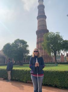 I am holding a 3D-printed model of the Qutub MInar while standing in front of the real Minar. The Minar is a high tower (minerette) in three sections that get thinner the higher up they are. 